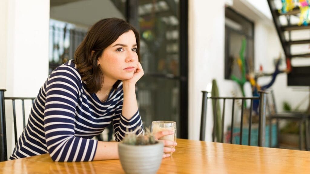 Woman in a striped shirt sits alone at a table with a drink, resting her head on her hand, expressing the isolation and frustration behind the question why can't I meet anyone