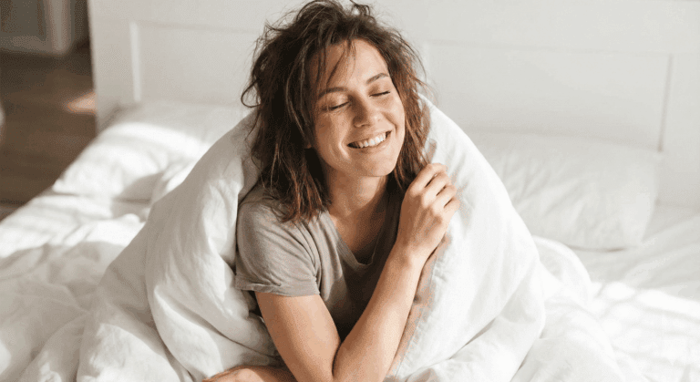 Smiling young woman sitting up in bed wrapped in white bedding, eyes closed, conveying contentment after self-pleasure.