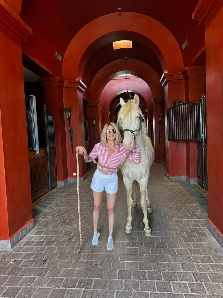 Dr. Sydney Ceruto standing with a purebred white Arabian horse inside the red-arched stables of Selman Marrakech, Morocco