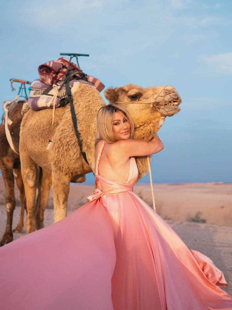 Dr. Sydney Ceruto in a flowing pink gown embracing a camel during a flying dress photo shoot in the Moroccan Sahara desert at sunset