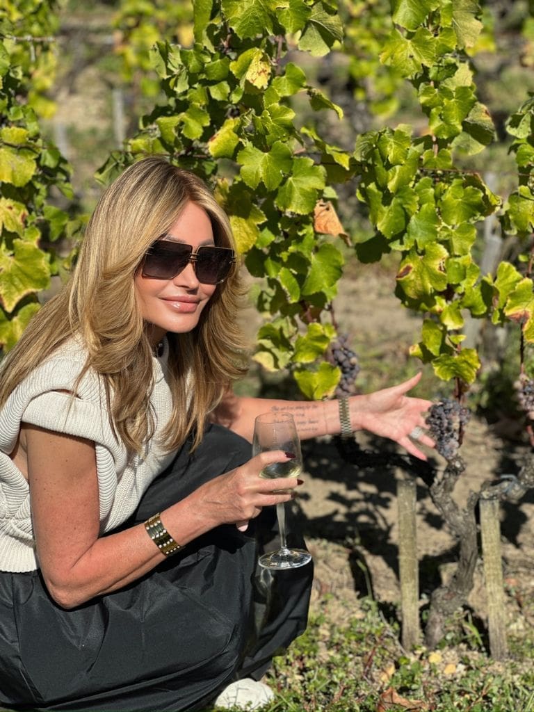 Dr. Sydney Ceruto holding a glass of wine while touching ripe grapes on the vine in a sunlit vineyard in Sauternes, France